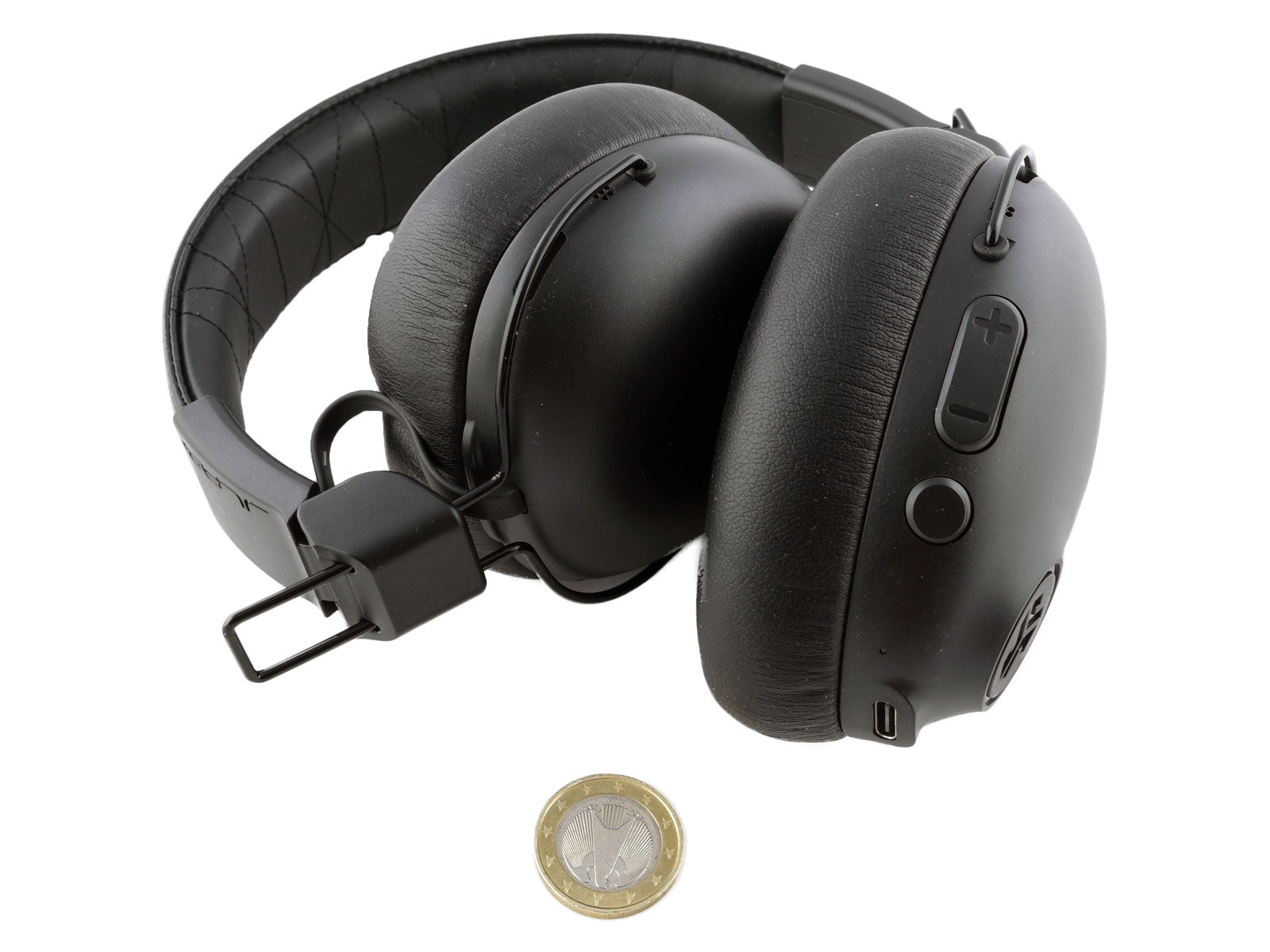 JLab Studio Pro ANC Wireless Over-Ear Kopfhörer Schwarze Ohrenhörer auf einer weißen Oberfläche, darunter eine kleine Münze als Maßstab.